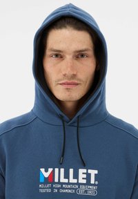 Niebieska bluza z kapturem i sznurkiem, z białym, czerwonym i niebieskim logo. Materiał wydaje się miękki; okolica szyi ma zaokrąglony kształt.