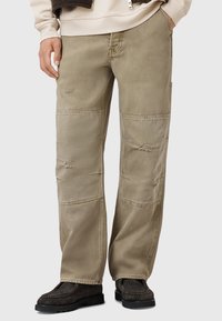 Beige cargo pants with a relaxed fit. Features distressed accents and patchwork details. Constructed from durable cotton blend material.