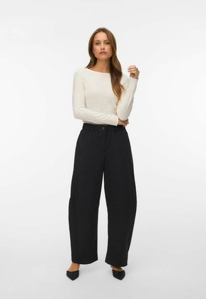 Femme debout, les bras croisés, portant un haut texturé blanc à manches longues, un pantalon large noir et des chaussures noires pointues.
