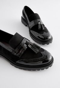 Schwarze Loafers aus einer Mischung aus Wildleder und Lackleder, verziert mit Brogue-Perforationen und dekorativen Quasten auf einer dicken Sohle.
