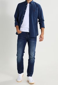 Marinblå skjorta med knappar över en vit T-shirt, kombinerad med mörkblå jeans och vita skor. Har en bröstficka och klassisk kragsdesign.