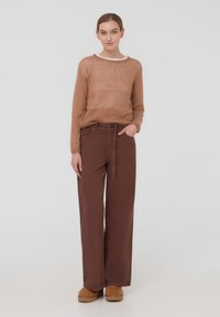 Maglione marrone lavorato a maglia con una vestibilità ampia e un tessuto a trama aperta abbinato a pantaloni marroni a vita alta e gamba larga. Le ciabatte marroni chiaro completano il look.