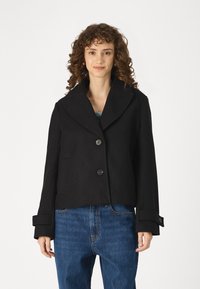 Black cropped jacket made of wool blend, featuring a wide collar, two large buttons, and roll-up cuffs, paired with blue high-waisted jeans.