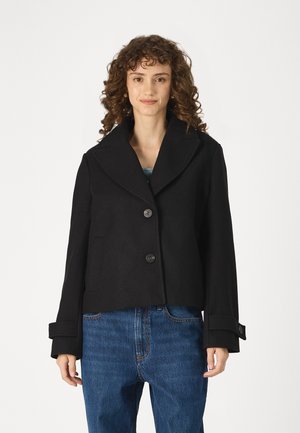 Svart cropped jakke laget av ullblanding, med bred krage, to store knapper og oppbrettede ermer, sammen med blå høytalede jeans.