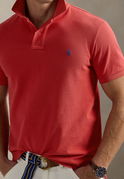 Bărbat purtând tricou polo roșu cu mâneci scurte și logo albastru, pantaloni albi, curea cu dungi albastre și galbene, și ceas de mână închis la culoare cu curea de piele.