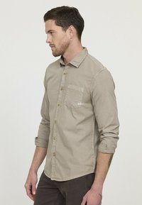 Chemise boutonnière beige clair avec une seule poche poitrine, manches longues retroussées et des détails de couture subtils. Tissu durable avec une texture lisse.