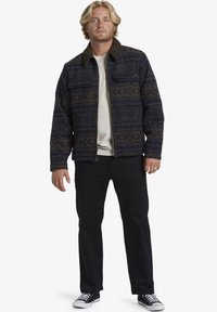 Navy jacket with geometric patterns and a brown fur collar, featuring a zip front, two chest pockets, and worn with black pants and sneakers.