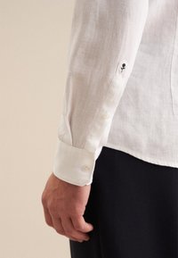 Camicia bianca in lino con polsini abbottonati, caratterizzata da un piccolo dettaglio ricamato a forma di rosa. Il tessuto è leggero, traspirante e con una texture particolare.