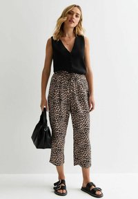 Zwarte mouwloze top met knopen, gecombineerd met lichtbruine broeken met luipaardprint. Zwarte sandalen en een bijpassende handtas maken de look compleet.