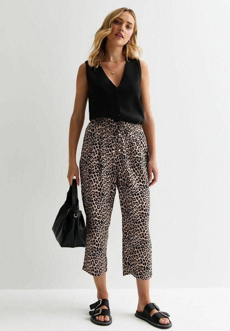 Zwarte mouwloze top met knopen, gecombineerd met lichtbruine broeken met luipaardprint. Zwarte sandalen en een bijpassende handtas maken de look compleet.