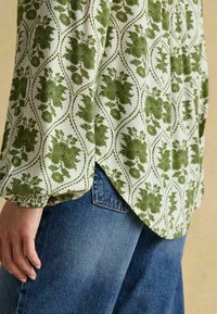 Zaļš ziedu raksts uz balta fona, ar garām piedurknēm un elastīgām aprocēm, kombinēts ar ziliem džinsiem.