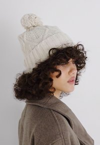 Beanie lavorato a maglia in beige chiaro con motivi testurizzati e un pompon in cima. Presenta un materiale morbido e accogliente e una vestibilità aderente.