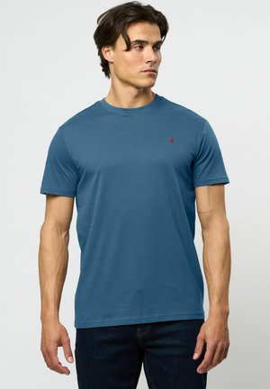 Camiseta de algodón azul con cuello redondo y mangas cortas. Presenta un pequeño logo rojo en el lado izquierdo del pecho. Se lleva con jeans oscuros.