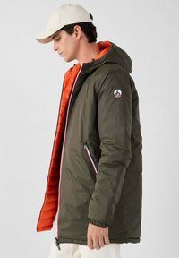 Giacca puffer verde oliva con interno arancione, dotata di cappuccio, tasche con zip e una toppa con logo sulla manica.