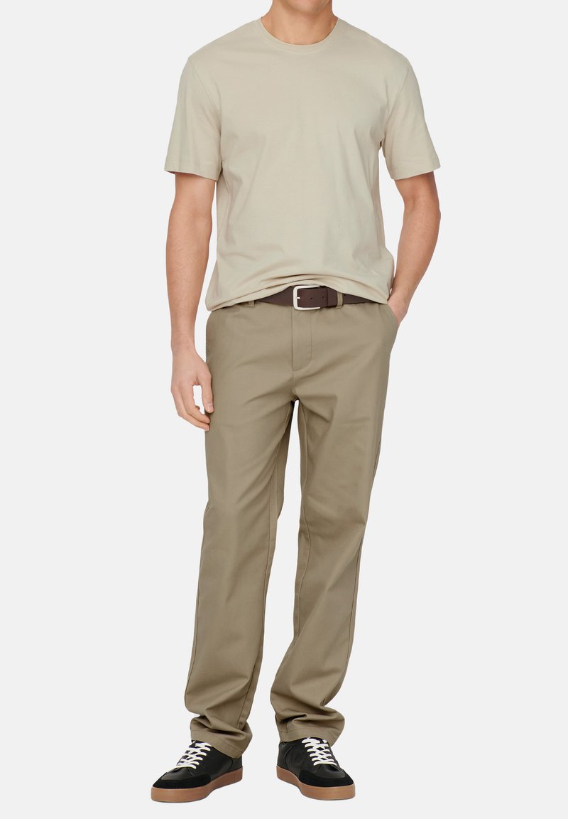 Beige t-shirt met korte mouwen, gedragen met lichte khaki broek, een bruine riem en zwarte sneakers met witte veters en bruine zolen.