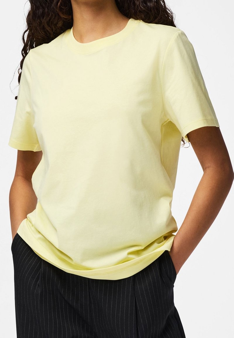 Femme portant un t-shirt jaune pastel à manches courtes et un pantalon noir à fines rayures, debout les mains dans les poches devant un fond uni.