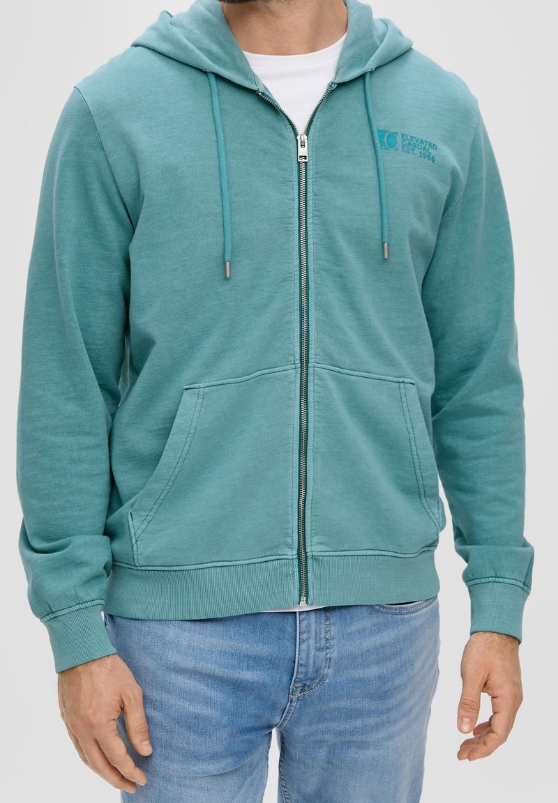 Homme portant un sweat à capuche zippé bleu sarcelle avec des poches avant et un jean bleu clair, debout devant un fond uni.
