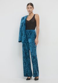 Pantaloni a palazzo in velluto turchese con una maglia nera a rete testurizzata. Il modello tiene in mano un blazer turchese abbinato. Tacchi a punta neri completano l'outfit.