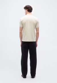 Άνδρας με την πλάτη στραμμένη φοράει μπεζ κοντομάνικο πόλο μπλουζάκι, μαύρα φαρδιά τζιν και μαύρα παπούτσια σε λευκό φόντο.