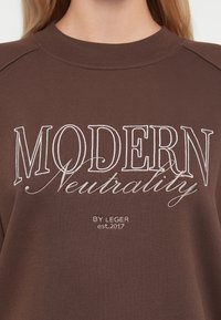 Sweatshirt marron en tissu doux, avec un texte brodé blanc "MODERN Neutrality" et le logo "BY LEGER est.2017" en dessous.