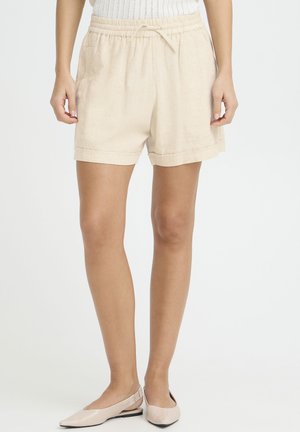 Kvinde iført beige linnedshorts med snøre og lysebeige spidse flade sko, stående mod en hvid baggrund.