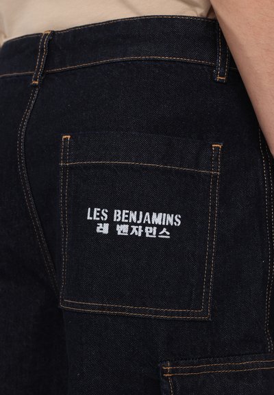 Pantalons en denim foncé avec des coutures visibles en orange, une poche arrière avec "LES BENJAMINS" brodé en blanc, et une finition en tissu texturé.