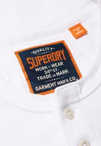 Bílá bavlněná košile s námořnickou a oranžovou etiketou, na které je napsáno "Superdry," "WORK-WEAR" a další text. Na límci jsou viditelné dva knoflíky.