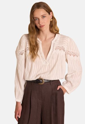 Blusa beige chiaro con sottili righe, dettagli in pizzo, scollo a V, maniche lunghe e tessuto texturizzato, abbinata a pantaloni marrone scuro.