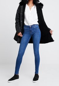 Abrigo negro de invierno con borde de piel, camisa blanca de botones, vaqueros ajustados azules y zapatos negros sin cordones. Texturas suaves y diseño casual.