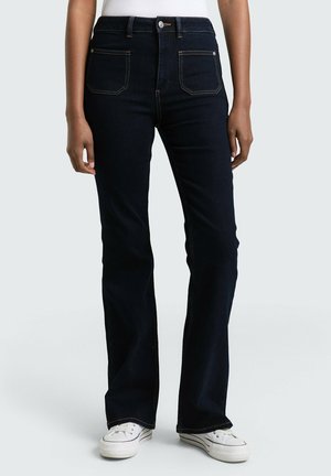 Osoba nosząca ciemnoniebieskie proste dżinsy z przednimi kieszeniami naszywanymi oraz białe trampki na jasnym tle.