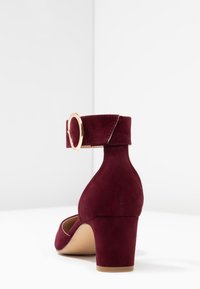 Burgundy mockaläder klackskor med rund tå och ankelsnodd, prydd med en guldfärgad, cirkulär spänne. Blockklackdesign med en slät yta.