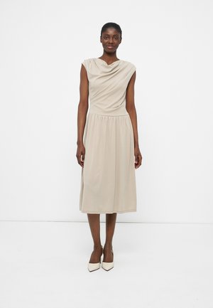 Moteris vilki beige spalvos midi suknelę be rankovių, su drapiruotu iškirpte, dėvi baltus batelius su smailiais nago galais, stovi prieš paprastą baltą foną.