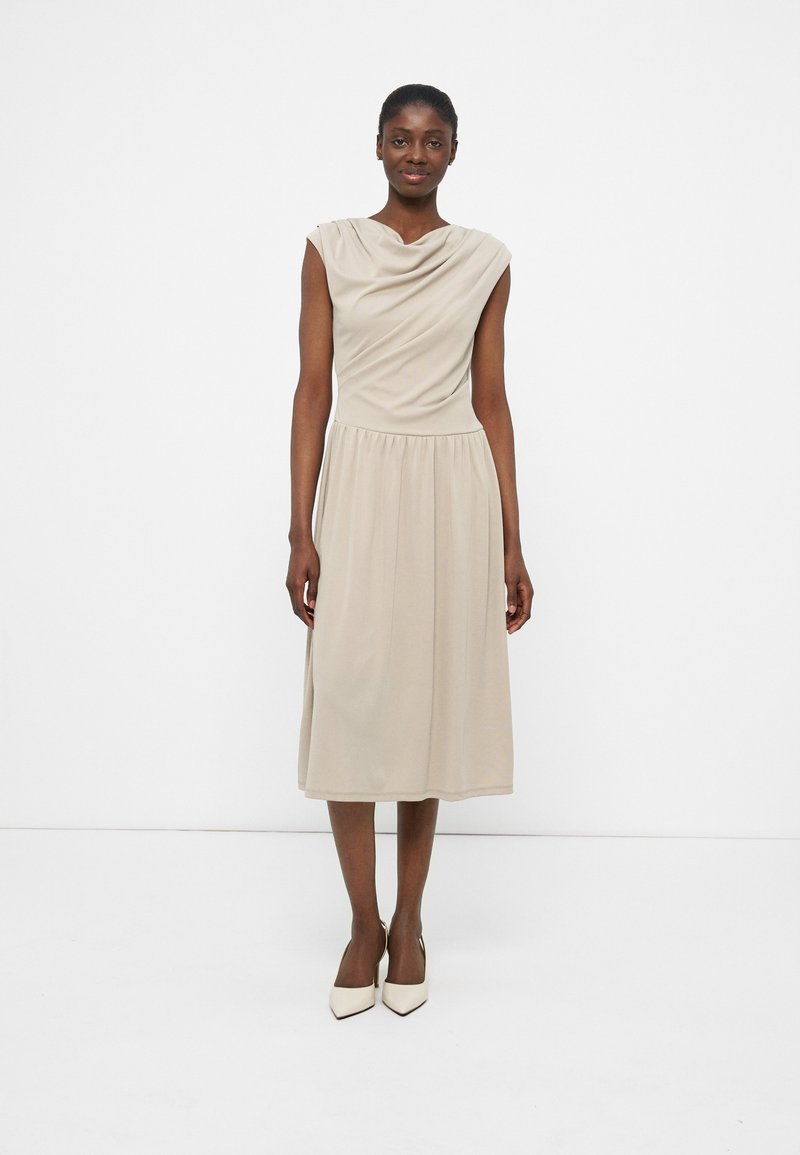 Γυναίκα που φοράει μπεζ μίντι φόρεμα χωρίς μανίκια με ντεκολτέ τύπου draped και λευκές γόβες μυτερές, στέκεται μπροστά σε απλό λευκό φόντο.