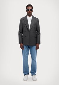 Blazer grigio doppiopetto con rever a punta, abbinato a una camicia bianca a righe, jeans azzurri chiari e sneakers bianche; accessorato con occhiali da sole.