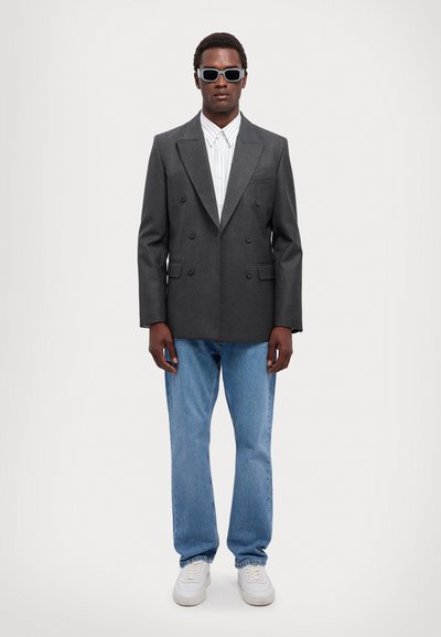 Grå dobbeltbrystet blazer med slidslapper, parret med en hvid stribet skjorte, lyseblå jeans og hvide sneakers; accessoriseret med solbriller.