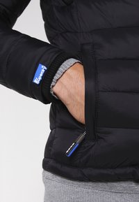 Sort pufferjakke med en quiltet design, der har en grå ribbet manchet og en mærket blå lap på armen. Inkluderer en tilgængelig lynlås-lomme.