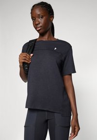 Ung kvinde med fletninger bærer en mørkeblå kortærmet skjorte og leggings, mens hun holder et sort rygsækerem på sin skulder.
