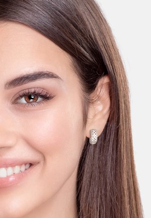 Silberne Creolen mit einem geflochtenen Gitterdesign. Die Ohrringe sind auf glatter, heller Haut platziert, wobei lange, dunkle Haare sanft das Gesicht umrahmen.