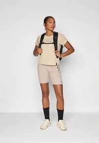 Nainen, jolla on päällä vaaleanbeige t-paita ja siihen sopivat shortsit, musta reppu, mustat sukat ja vaaleat lenkkarit, seisoo kädet repun hihnoilla.