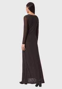 Langes braunes Kleid mit langen durchsichtigen Ärmeln und geradem Schnitt. Verfügt über einen U-förmigen Rückenausschnitt und einen Schlitz an der Seitennaht am Saum. Trägt schwarze Schuhe.