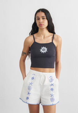 Νέα γυναίκα με μακριά σκούρα μαλλιά που φοράει μαύρο crop top με το λογότυπο "Santa Cruz" και λευκά σορτς διακοσμημένα με μοτίβα μπλε αστερία και κοχυλιών.