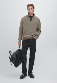 Mladý muž v béžovém svetru na zip a černých kalhotách, držící černý kožený batoh, stojící před světlým neutrálním pozadím.