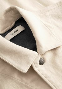 Close-up af en beige bomuldsjakke med krave, metaltrykknap og sort indvendig foring, der viser mærkeetiketten "GABBA".