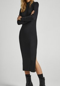 Černé žebrované pletené šaty s dlouhými rukávy, vysokým límcem, bočním rozparkem a upnutým siluetou, doplněné černými kozačkami po kolena.