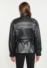 Giacca di pelle nera con vita cinturata, colletto alto, dettagli con bottoni sulle spalle e una texture liscia e lucida.