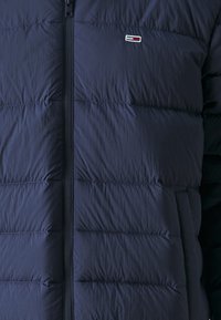 Marineblauwe gewatteerde jas met ritssluiting aan de voorkant, doorgestikt ontwerp en een klein geborduurd logo. Het materiaal is glad met een zachte glans en geïsoleerde textuur.