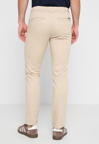 Bărbat îmbrăcat cu pantaloni slim-fit bej și adidași maro de la Adidas, stând cu spatele la cameră pe un fundal simplu deschis.
