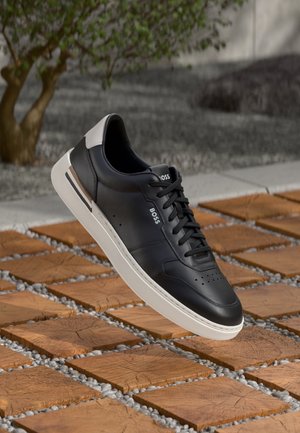Czarny skórzany sneaker BOSS z białą podeszwą i detalem na pięcie, przedstawiony unoszący się nad wzorzystą, drewniano-kamienną nawierzchnią na zewnątrz.