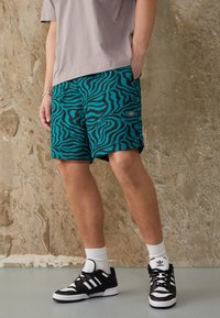 Shorts de bain turquoise avec un motif rayé zèbre noir, fabriqués en matériau léger. Comprend des poches latérales et un détail en patch avec logo.