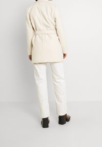 Cappotto lungo beige con cintura, design con colletto e silhouette elegante, abbinato a pantaloni bianchi a vita alta e tacchi neri spessi.
